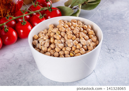 White bowl filled with chickpeas beside cherry tomatoes and bay leaves on textured surface 141328065
