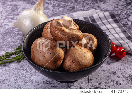 Fresh brown mushrooms in black bowl with garlic rosemary and cherry tomatoes on textured surface Fresh brown mushrooms in black bowl with garlic rosemary and cherry tomatoes on textured surface 141328242