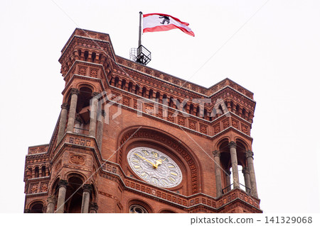 Berlin red town hall with Berlin flag flying in the cloudy sky. Detailed view of Rotes Rathaus clock tower, iconic landmark featuring ornate brickwork representing the city history. Copy space Berlin red town hall with Berlin flag flying in the cloudy sky. Detailed view of Rotes Rathaus clock tower, iconic landmark featuring ornate brickwork representing the city history. Copy space 141329068