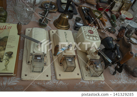 XALO, SPAIN - JUNE 20, 2026.Three vintage mechanical wall mounted coffee grinders sit on a table. They are next to other antique kitchen items at a flea market. People browse and shop for collectibles XALO, SPAIN - JUNE 20, 2026.Three vintage mechanical wall mounted coffee grinders sit on a table. They are next to other antique kitchen items at a flea market. People browse and shop for collectibles 141329285