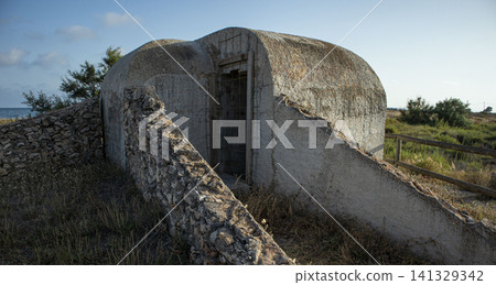 A concrete bunker from the Civil War sits in the salt marshes of Santa Pola, Alicante, Spain. It shows signs of wear and age, surrounded by grass and a stone wall. A concrete bunker from the Civil War sits in the salt marshes of Santa Pola, Alicante, Spain. It shows signs of wear and age, surrounded by grass and a stone wall. 141329342