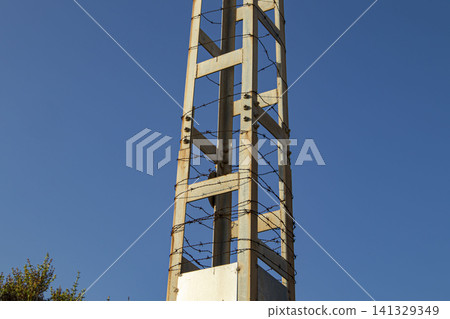 A tall utility tower stands close up, featuring rustic metal lattice and wrapped in barbed wire under a clear blue sky during the day. 141329349