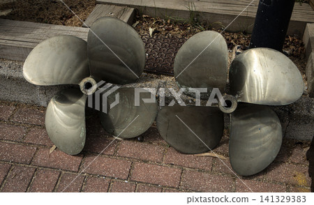 A pair of heavy brass boat propellers with green patina sit on brick pavement at a flea market. Sunlight shines on them as people walk by browsing various items. A pair of heavy brass boat propellers with green patina sit on brick pavement at a flea market. Sunlight shines on them as people walk by browsing various items. 141329383