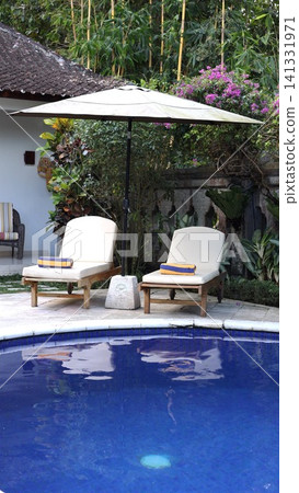 Poolside lounge chairs beneath large patio umbrella beside blue tiled swimming pool in tropical Bali garden with bamboo, bougainvillea, stone wall, and tranquil resort setting Poolside lounge chairs beneath large patio umbrella beside blue tiled swimming pool in tropical Bali garden with bamboo, bougainvillea, stone wall, and tranquil resort setting 141331971