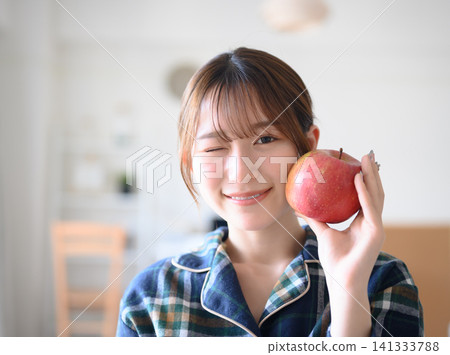 一位身穿家居服的女子一邊眨眼一邊拿著蘋果。 一位身穿家居服的女子一邊眨眼一邊拿著蘋果。 141333788