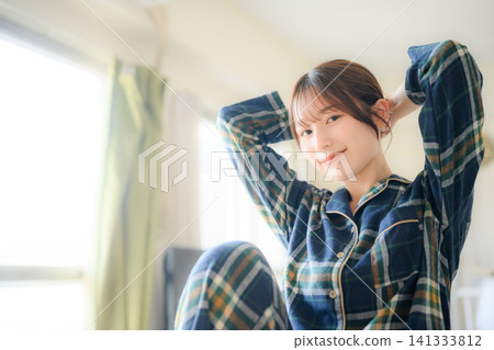 A woman in pajamas is fixing her hair in her room. 141333812