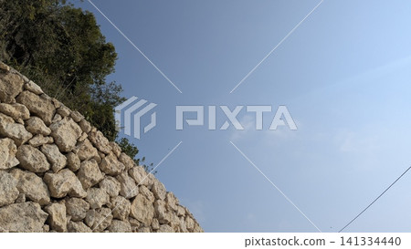 A horizontal wide angle perspective captures a curved dry stone wall built from weathered limestone blocks below a clear light blue sky. Green tree foliage emerges from behind the stony structure, A horizontal wide angle perspective captures a curved dry stone wall built from weathered limestone blocks below a clear light blue sky. Green tree foliage emerges from behind the stony structure, 141334440
