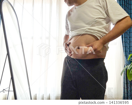 A middle-aged obese man pinches his stomach while looking in the mirror. 141336595