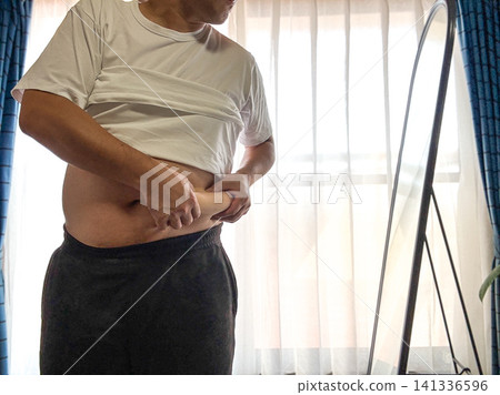 一位中年男子照鏡子時,對自己的腹部脂肪感到擔憂。 一位中年男子照鏡子時,對自己的腹部脂肪感到擔憂。 141336596