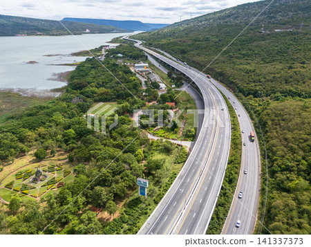 Drone aerial view of a highway running beside a reservoir with lush green mountains, Transportation infrastructure and renewable energy concept Drone aerial view of a highway running beside a reservoir with lush green mountains, Transportation infrastructure and renewable energy concept 141337373