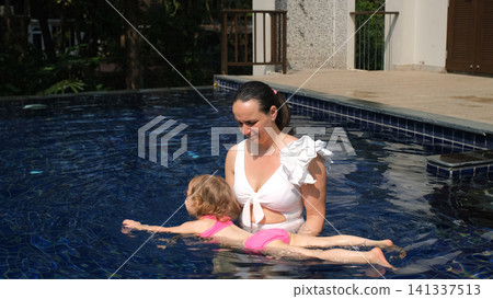 Young mother in white swimsuit teaching her small daughter to swim and float in a blue resort swimming pool. Swimming lesson concept 141337513
