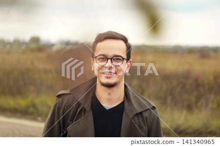 Portrait of handsome young man with glasses smiling outdoors in autumn field. Happy confident caucasian male looking at camera wearing stylish coat, autumn season concept Portrait of handsome young man with glasses smiling outdoors in autumn field. Happy confident caucasian male looking at camera wearing stylish coat, autumn season concept 141340965