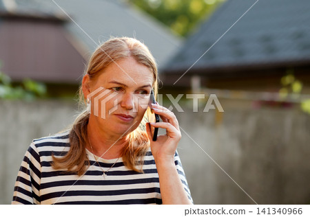 Middle-aged woman talking on smartphone outdoors in backyard. Mature caucasian female in striped t-shirt having phone conversation, listening carefully with thoughtful expression, communication Middle-aged woman talking on smartphone outdoors in backyard. Mature caucasian female in striped t-shirt having phone conversation, listening carefully with thoughtful expression, communication 141340966