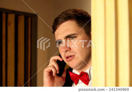 Confused or worried man in a suit and red bow tie talking on a smartphone, looking away indoors. Portrait of worried groom in bowtie talking on smartphone and peeking from behind a wall Confused or worried man in a suit and red bow tie talking on a smartphone, looking away indoors. Portrait of worried groom in bowtie talking on smartphone and peeking from behind a wall 141340969
