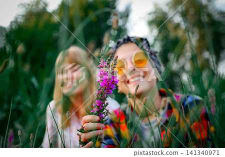 Smiling young friends in a green field holding a purple wildflower, with one wearing yellow round sunglasses. Two Happy Young Women Friends Laughing with Purple Flower in a Summer Nature Field Smiling young friends in a green field holding a purple wildflower, with one wearing yellow round sunglasses. Two Happy Young Women Friends Laughing with Purple Flower in a Summer Nature Field 141340971