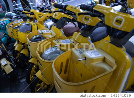A perspective close-up shot of vibrant yellow electric rental scooters with front baskets neatly lined up on an urban city sidewalk. A perspective close-up shot of vibrant yellow electric rental scooters with front baskets neatly lined up on an urban city sidewalk. 141341059