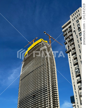 Tall curved high-rise building under construction with cranes and clear blue sky. Tall curved high-rise building under construction with cranes and clear blue sky. 141341519