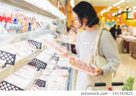 A woman in her 50s holding two meat packs in the supermarket, struggling to decide which one to buy. A woman in her 50s holding two meat packs in the supermarket, struggling to decide which one to buy. 141343825