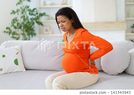 Black pregnant woman leaning forward on sofa and holding her lower back Black pregnant woman leaning forward on sofa and holding her lower back 141343943