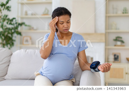 Black pregnant woman checking her blood pressure at home while touching temple Black pregnant woman checking her blood pressure at home while touching temple 141343963