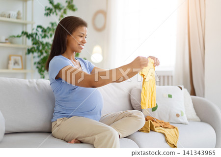 Black pregnant woman sitting on sofa and holding up yellow baby garment Black pregnant woman sitting on sofa and holding up yellow baby garment 141343965
