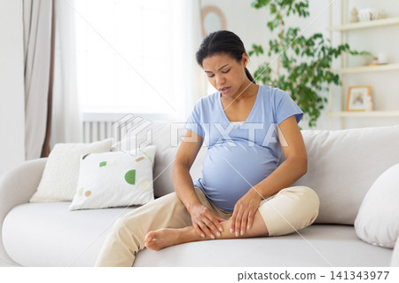 Black pregnant woman sitting on sofa and massaging ankle, showing leg pain Black pregnant woman sitting on sofa and massaging ankle, showing leg pain 141343977