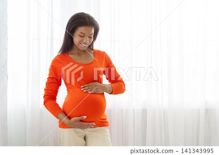 Smiling Black pregnant woman standing by bright window and cradling her belly 141343995