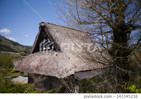 A thatched roofed house with a blue sky and mountain range as a backdrop; decorative design of the gable wall. A thatched roofed house with a blue sky and mountain range as a backdrop; decorative design of the gable wall. 141345238