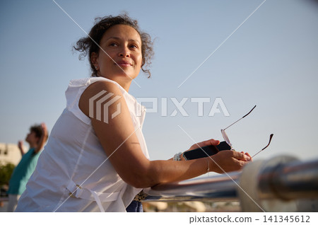 Smiling woman leaning on railing outdoors holding sunglasses and phone on a sunny summer day Smiling woman leaning on railing outdoors holding sunglasses and phone on a sunny summer day 141345612