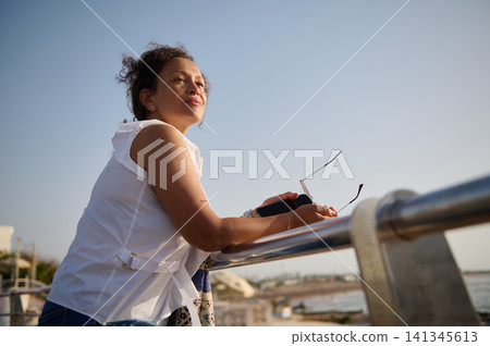 Woman leaning on waterfront railing holding sunglasses and phone, looking relaxed at sunset Woman leaning on waterfront railing holding sunglasses and phone, looking relaxed at sunset 141345613