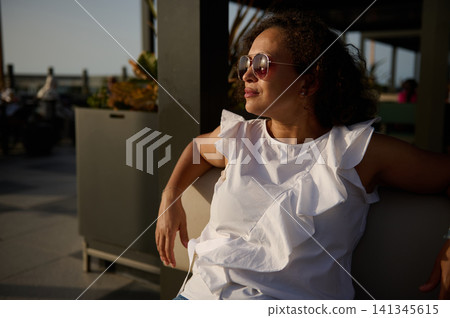 Woman relaxing on outdoor patio at sunset wearing sunglasses and white ruffled top Woman relaxing on outdoor patio at sunset wearing sunglasses and white ruffled top 141345615