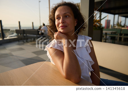 Thoughtful woman portrait at outdoor cafe during golden hour sunlight, relaxed summer evening Thoughtful woman portrait at outdoor cafe during golden hour sunlight, relaxed summer evening 141345618