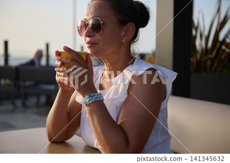 Woman in sunglasses enjoying morning coffee on a seaside cafe terrace, relaxed and thoughtful Woman in sunglasses enjoying morning coffee on a seaside cafe terrace, relaxed and thoughtful 141345632