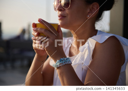 Woman in sunglasses holding a yellow coffee cup, relaxing outdoors at sunset with bracelets and ring Woman in sunglasses holding a yellow coffee cup, relaxing outdoors at sunset with bracelets and ring 141345633