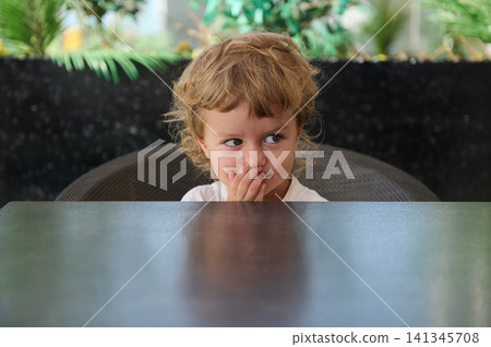 Shy toddler peeking over table in outdoor cafe with thoughtful, playful expression Shy toddler peeking over table in outdoor cafe with thoughtful, playful expression 141345708