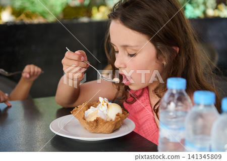 Young girl enjoying ice-cream in a waffle bowl while eating with a spoon at a cafe 141345809