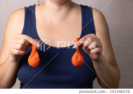Woman holding two deflated orange balloons over chest in navy tank top against neutral background, suggesting body measurement, weight change, wellness, self image, and personal health concept 141346258