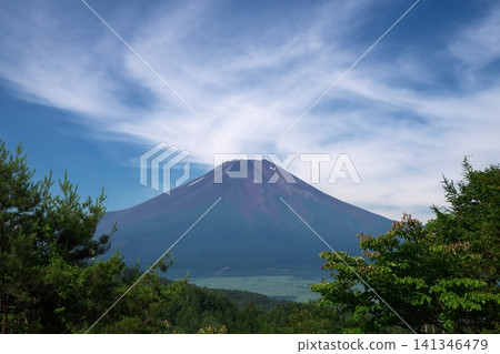 Mount Fuji in summer, as seen from Nijumagari Pass in Oshino Village. A natural landscape surrounded by dynamic clouds spreading across a blue sky and lush green trees. Mount Fuji in summer, as seen from Nijumagari Pass in Oshino Village. A natural landscape surrounded by dynamic clouds spreading across a blue sky and lush green trees. 141346479