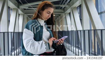 Young woman offering friendly glance in pedestrian corridor, slight smile while holding phone and bag, confident posture with relaxed shoulders, modern geometric background and soft daylight creating Young woman offering friendly glance in pedestrian corridor, slight smile while holding phone and bag, confident posture with relaxed shoulders, modern geometric background and soft daylight creating 141348197