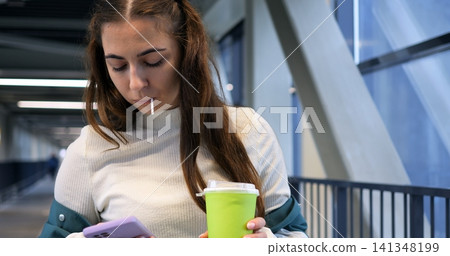 Woman browsing phone. Casual woman with coffee. Young woman relaxing and checking messages on campus bridge. Everyday campus scene with woman casually browsing messages while holding cup Woman browsing phone. Casual woman with coffee. Young woman relaxing and checking messages on campus bridge. Everyday campus scene with woman casually browsing messages while holding cup 141348199