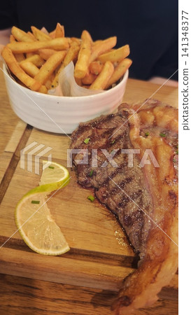 Grilled steak accompanied by crispy French fries and lemon wedges, presented on a wooden cutting board in a casual dining environment Grilled steak accompanied by crispy French fries and lemon wedges, presented on a wooden cutting board in a casual dining environment 141348377