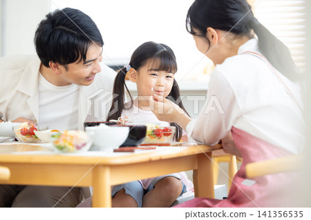 家庭用餐場景:一個小女孩臉頰沾著米粒。 家庭用餐場景:一個小女孩臉頰沾著米粒。 141356535