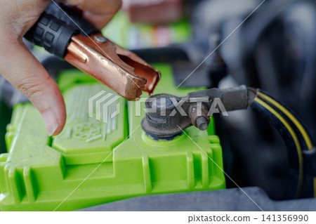 Hand holding jumper cable clamp near car battery Hand holding jumper cable clamp near car battery 141356990