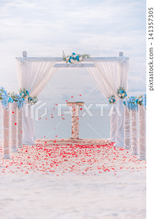 White wedding arch decorated with blue flowers and rose petals on tropical beach, vertical frame. Elegant destination wedding setup with sheer curtains on white sand by the sea. Romantic ceremony White wedding arch decorated with blue flowers and rose petals on tropical beach, vertical frame. Elegant destination wedding setup with sheer curtains on white sand by the sea. Romantic ceremony 141357305
