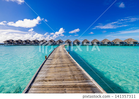 Wooden boardwalk perspective toward water villas in bright turquoise lagoon. Weathered planks leading to overwater suites under vivid sky with clouds, sunny resort day. Inviting luxury travel scene Wooden boardwalk perspective toward water villas in bright turquoise lagoon. Weathered planks leading to overwater suites under vivid sky with clouds, sunny resort day. Inviting luxury travel scene 141357311