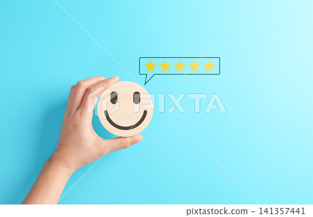 Customer satisfaction concept. Hand holding a wooden smiley face icon with a five-star rating speech bubble on blue background. Positive feedback and user experience evaluation. service excellence, Customer satisfaction concept. Hand holding a wooden smiley face icon with a five-star rating speech bubble on blue background. Positive feedback and user experience evaluation. service excellence, 141357441
