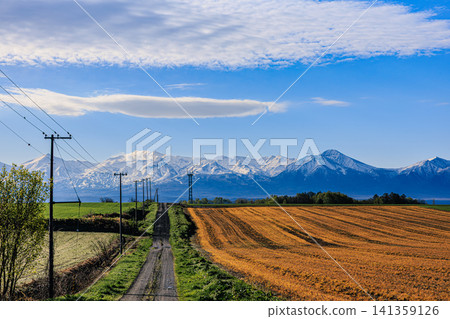 A single country road stretches towards the Tokachi mountain range, still covered in snow, against a backdrop of a beautiful blue sky, and vast fields of cultivated land. A single country road stretches towards the Tokachi mountain range, still covered in snow, against a backdrop of a beautiful blue sky, and vast fields of cultivated land. 141359126