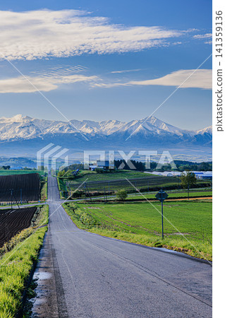 A landscape of snow-capped Tokachi mountain ranges and a single road stretching straight into a vast rolling hilly area. A landscape of snow-capped Tokachi mountain ranges and a single road stretching straight into a vast rolling hilly area. 141359136