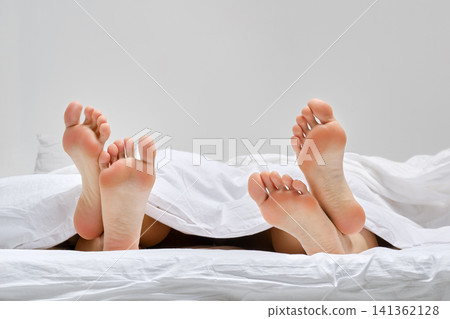 Two pairs of female feet on bed under blanket 141362128