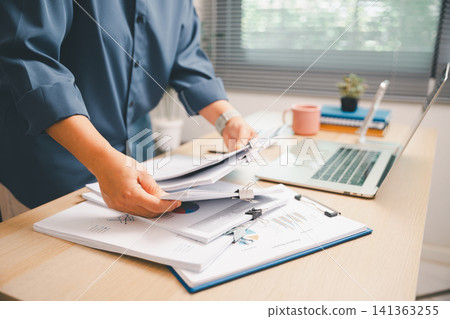 Hands organizing financial documents in a binder beside laptop and coffee on a bright office desk, showing accounting workflow, business report review, paperwork management, Hands organizing financial documents in a binder beside laptop and coffee on a bright office desk, showing accounting workflow, business report review, paperwork management, 141363255